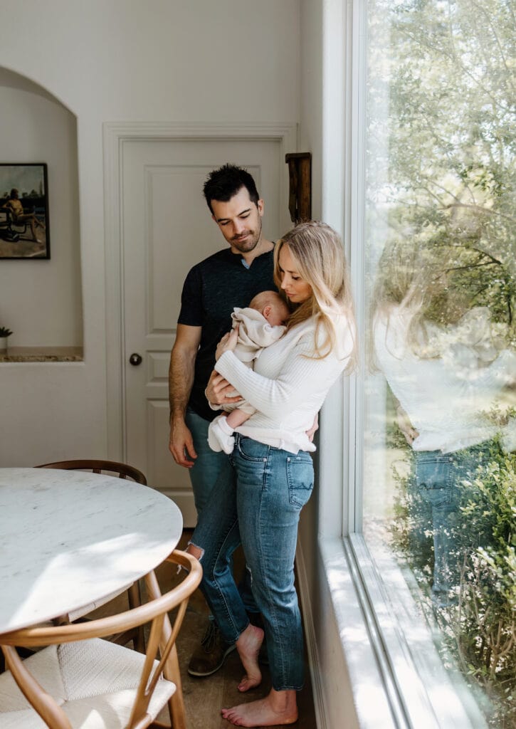 Austin Newborn Photographer, mother and father standing with newborn baby next to large window