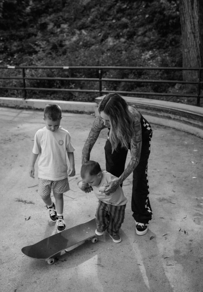 Black and white photo of a mother helping her two young sons with a skateboard.
