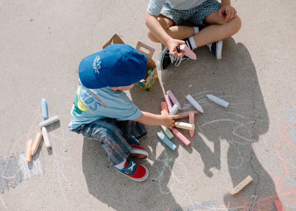 Two young brothers sitting on concrete, playing with colorful sidewalk chalk during a sunny afternoon outdoors.