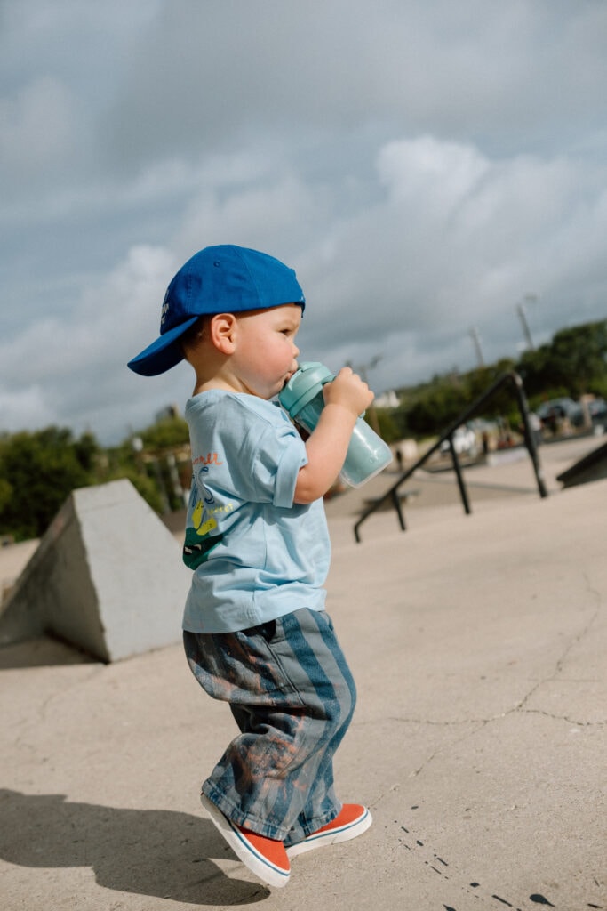 Young boy in a blue baseball cap drinking from a water bottle at an outdoor skatepark.