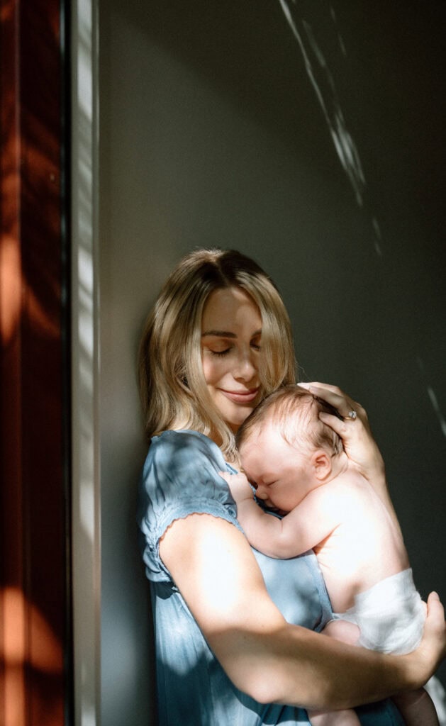 Mom in a blue dress snuggles her baby close in a patch of warm sunlight.