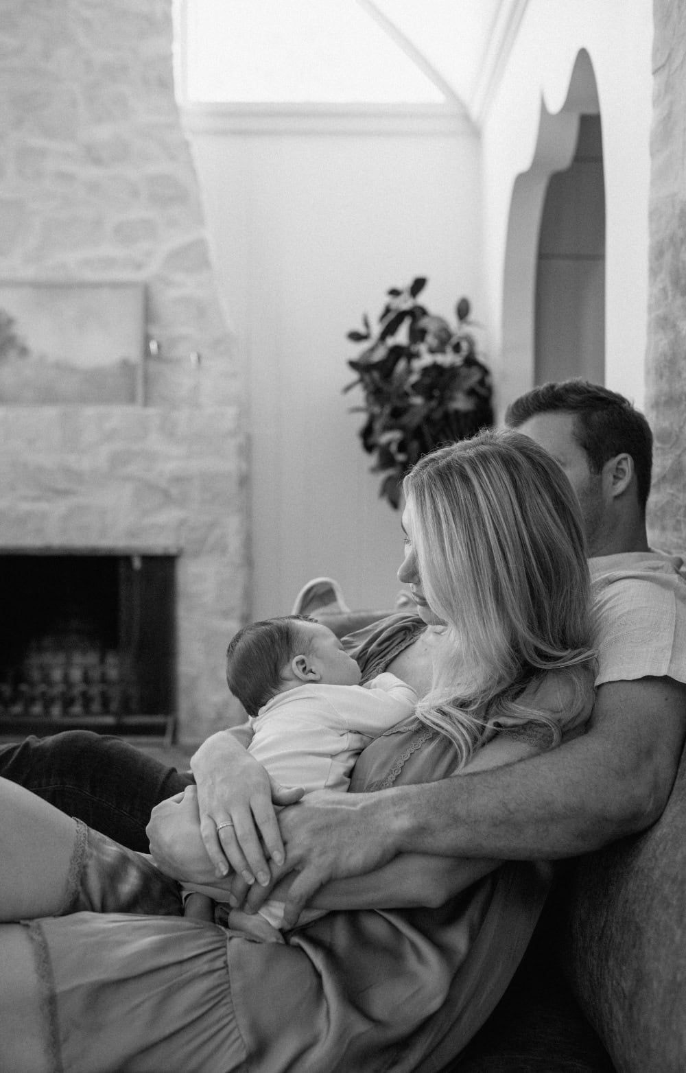 Black and white Austin newborn photography of parents cuddling their baby beside a stone fireplace.