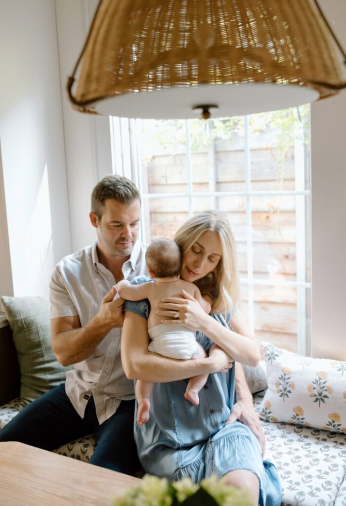 Mom hugs baby on a cozy window seat while dad gently rubs baby’s back.