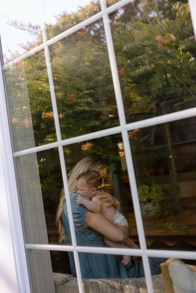 Leafy trees reflect in the window glass as mom holds her baby close inside.