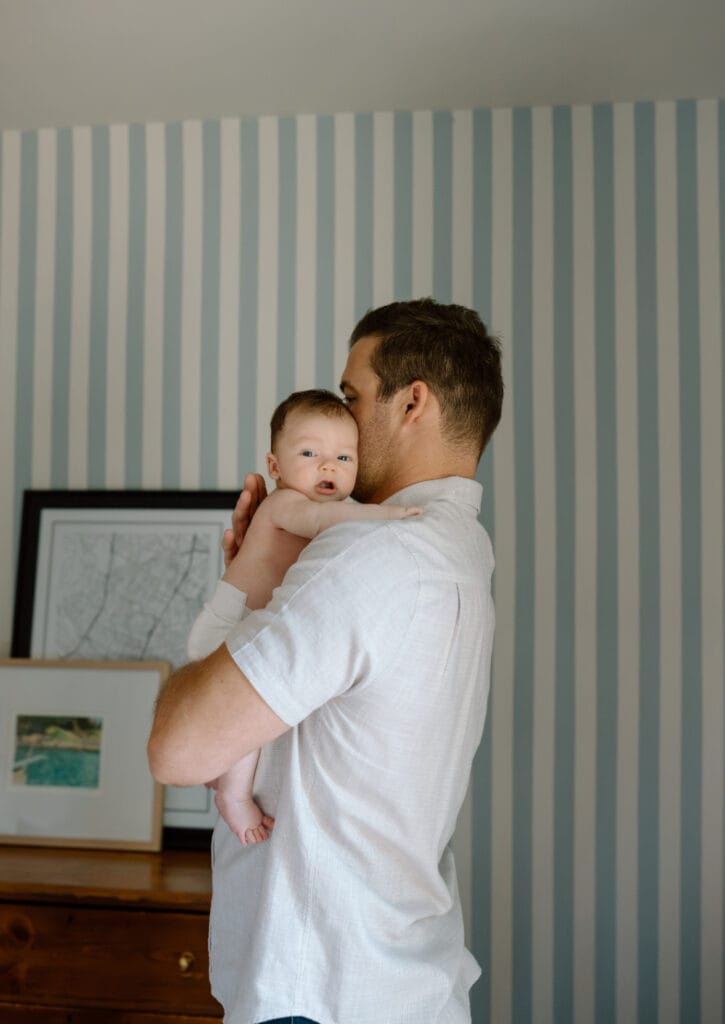 Dad kisses his baby’s head in a blue striped room during an Austin newborn photography session.