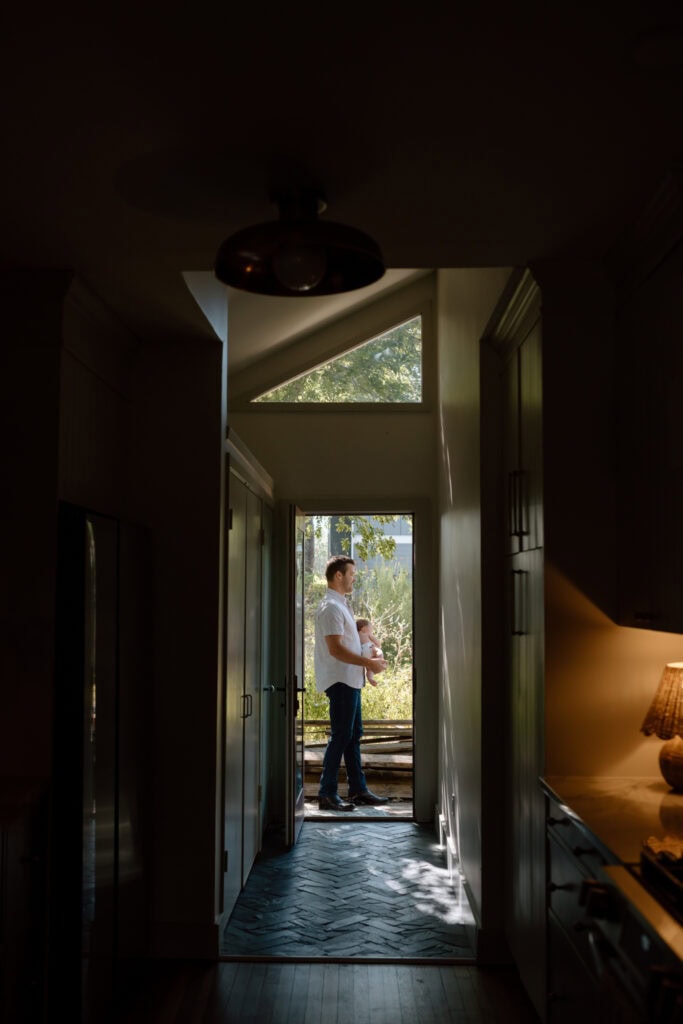 Dad holds his baby in a sunny open doorway, seen from down a dark hallway.