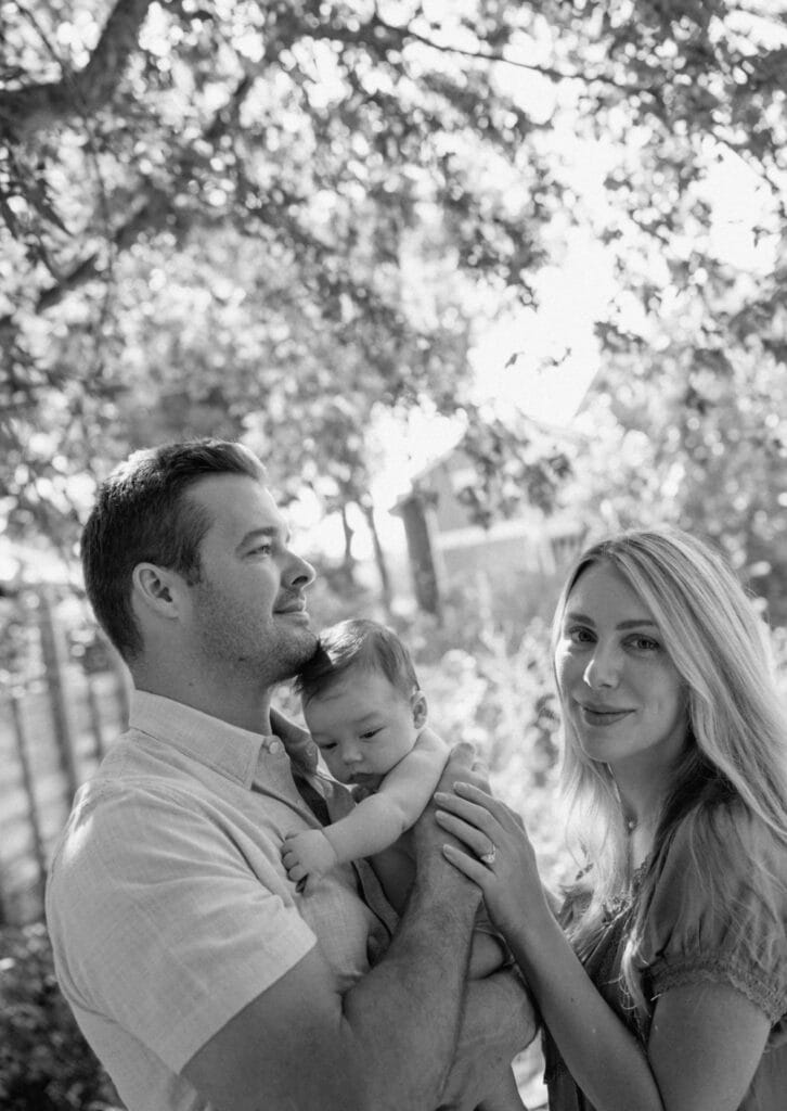 Mom smiles beside dad as he holds their baby close outside under sunny, leafy trees.